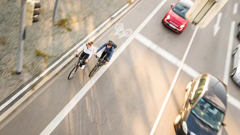 Zwei Fahrradfahrer:innen im Straßenverkehr mit Autos, von schräg vorne/oben fotografiert Zwei Fahrradfahrer:innen im Straßenverkehr mit Autos, von schräg vorne/oben fotografiert