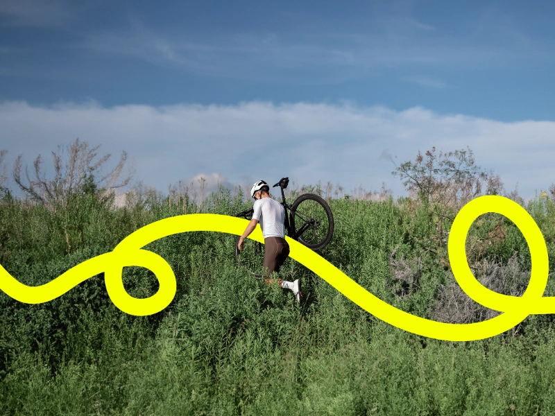Radler trägt sein Gravelbike durchs Gestrüpp, eine gelbe JobRad Loop Linie kringelt sich durchs Bild